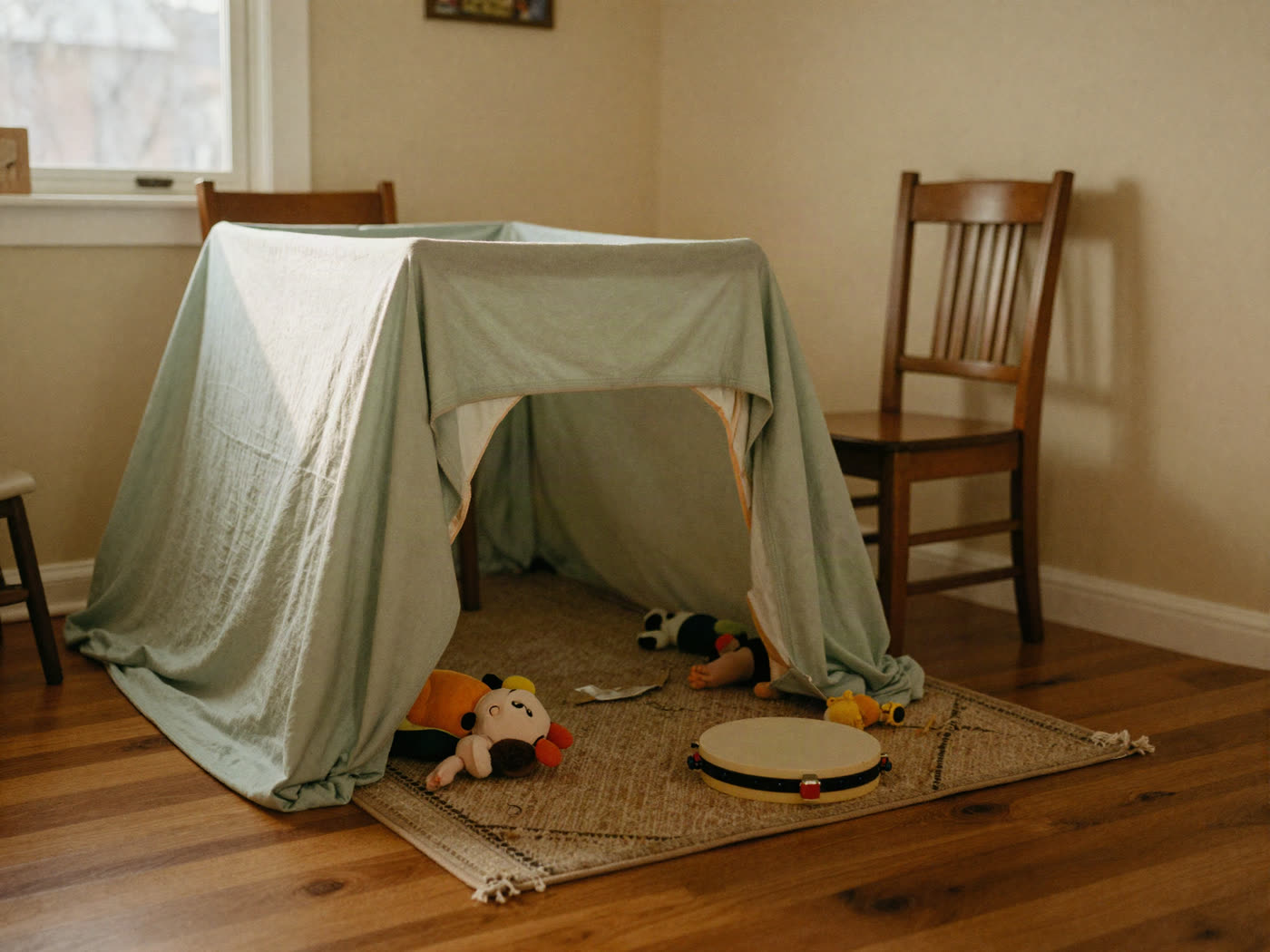 A bedsheet fort over two dining chairs with afternoon light coming through it and toys on the rug.